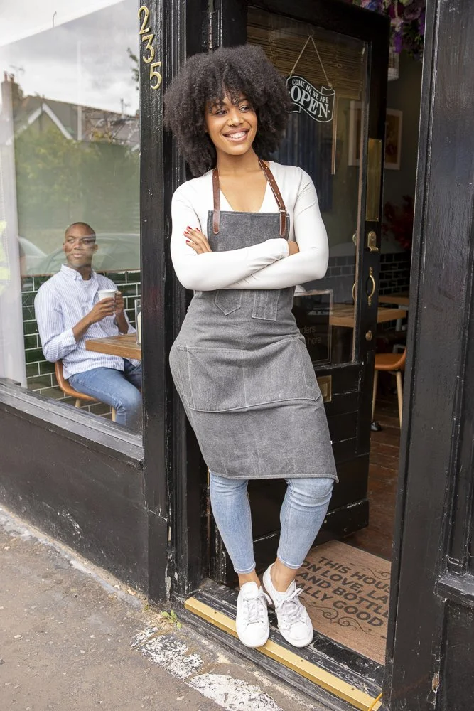 Smiling woman with curly hair wearing apron and white shirt standing at the entrance of a cafe, with another person sitting inside holding a drink.