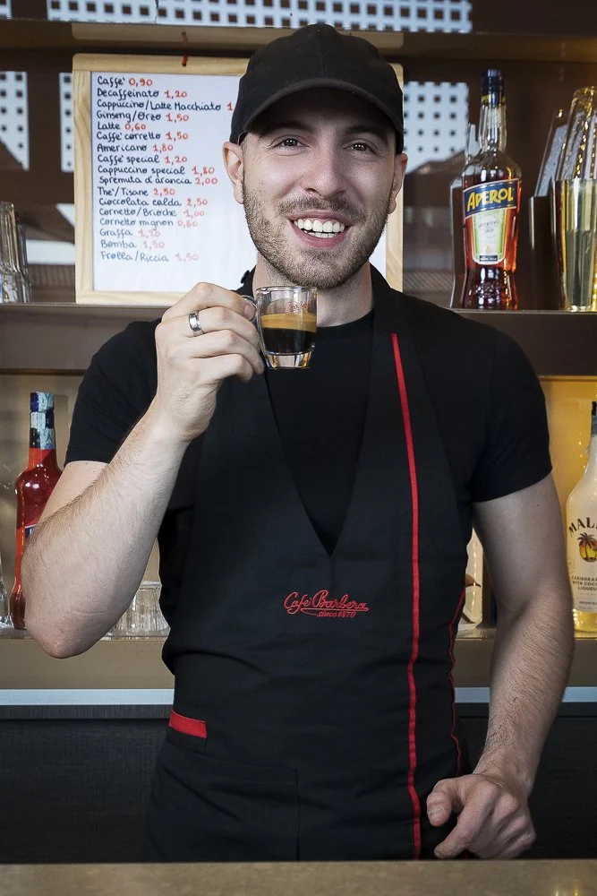 Smiling man in black apron holding an espresso shot at a coffee shop, behind a menu board and bottles of alcohol.