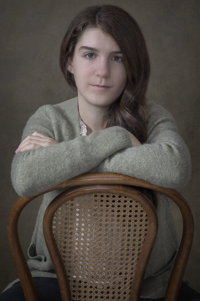 Portrait of a young woman with long brown hair, wearing a gray sweater, sitting with her arms crossed on a wooden chair with a woven back, against a plain dark background.