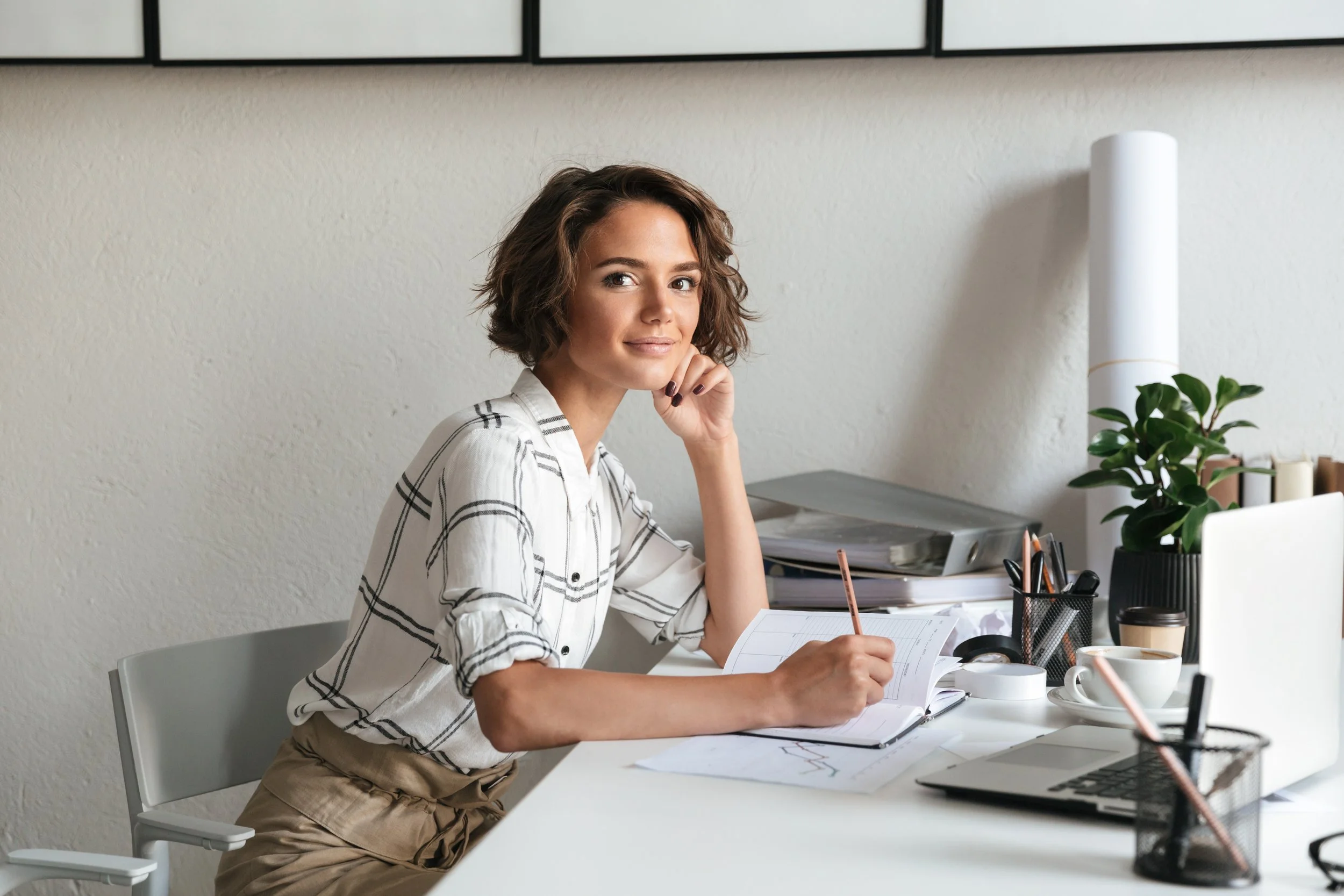 A woman sitting at a desk, writing in a notebook, with a laptop, coffee, and office supplies on the desk in a modern workspace.