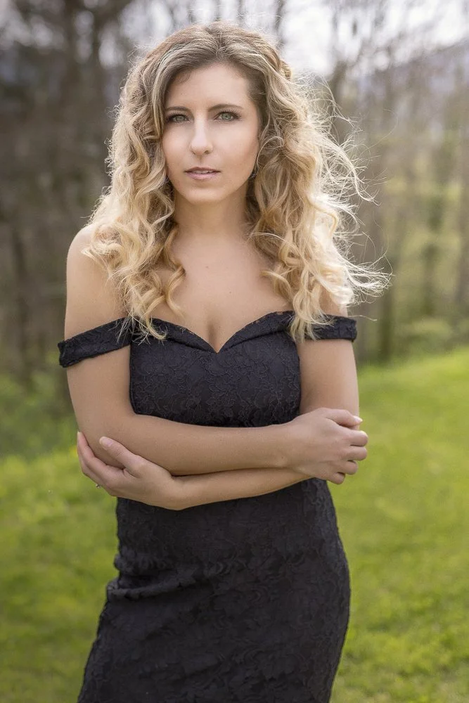 A woman with blonde curly hair wearing a black off-the-shoulder lace dress standing outdoors in a park with green grass and trees in the background.
