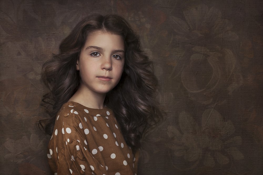 Portrait of a young girl with long wavy brown hair wearing a brown polka-dot shirt, standing against a textured dark background.