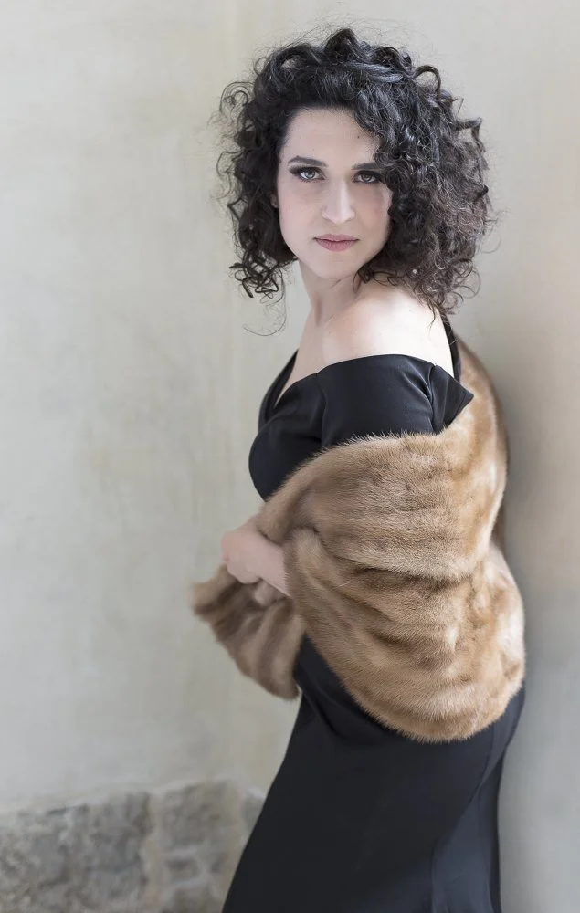 A woman with curly dark hair in a black off-shoulder dress wearing a brown fur stole, standing against a plain wall.