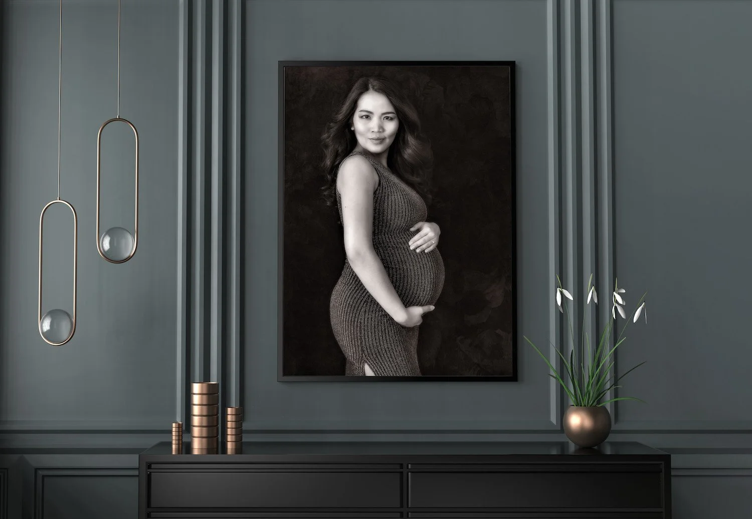 Black and white portrait of a pregnant woman in a form-fitting dress, displayed in a modern interior with gray walls, hanging pendant lights, and potted plants.