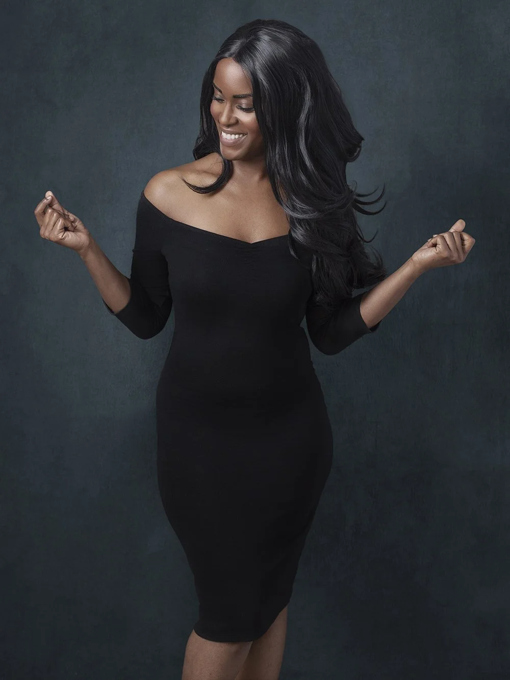A woman in a black off-shoulder dress smiling and posing snapping her fingers in celebration or excitement.