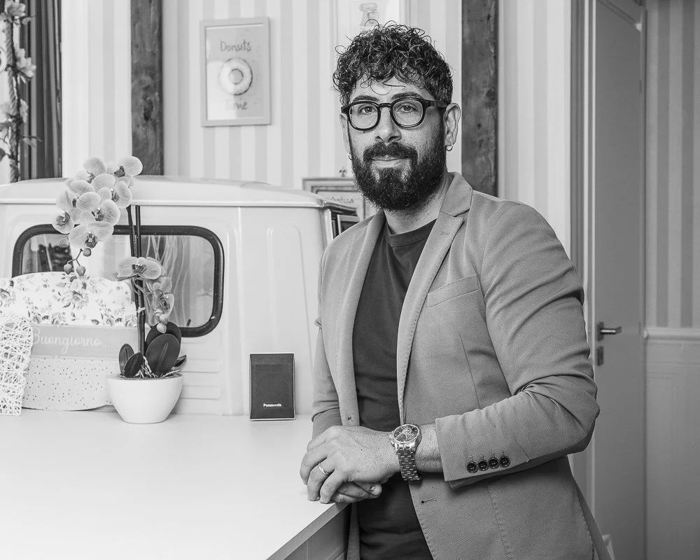 A man with curly hair and a beard wearing glasses, a blazer, and a watch, standing in a room with a large white van behind him, decorated with flowers and plants.