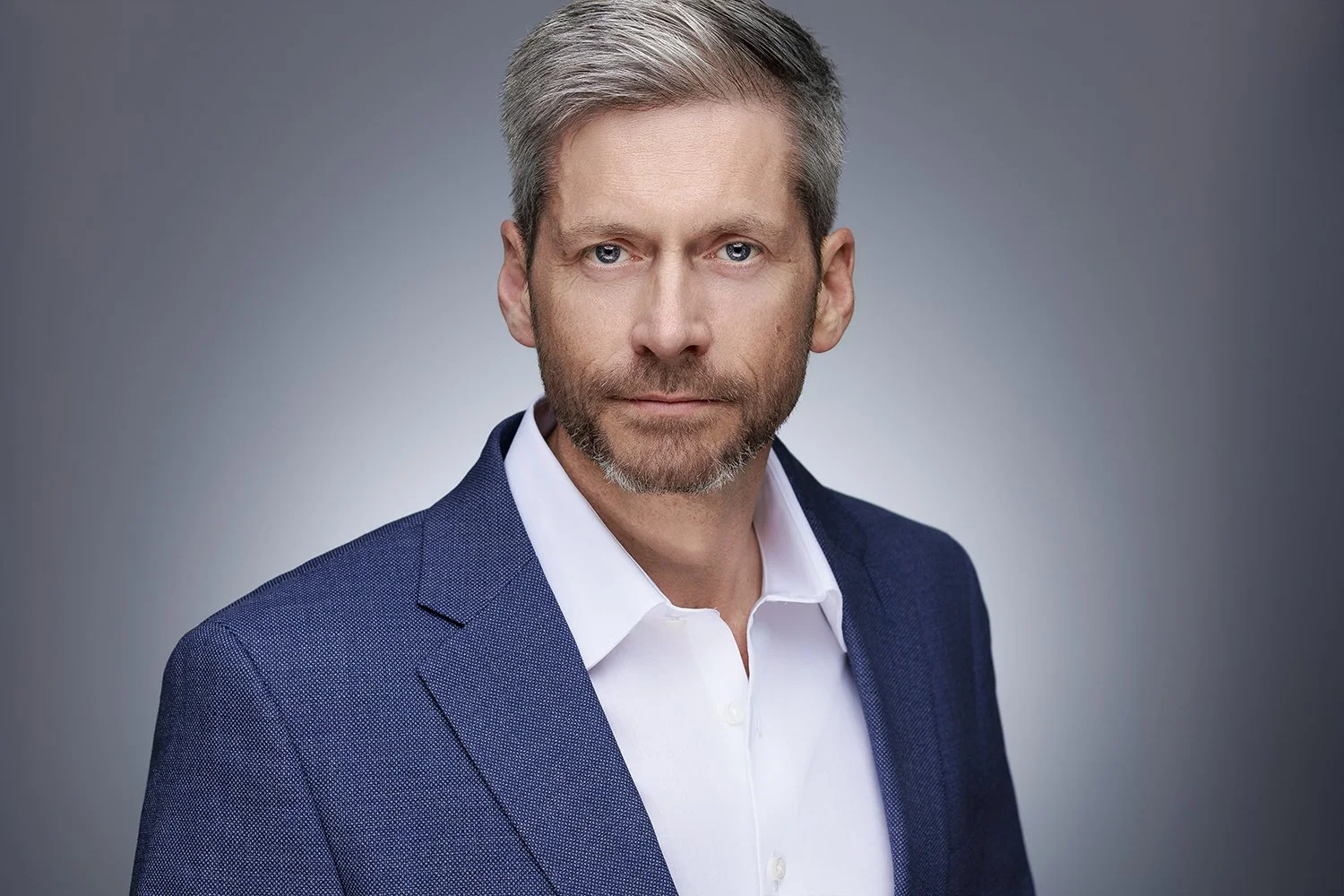 A portrait of a handsome man with gray hair and a beard, wearing a blue suit jacket over a white dress shirt, against a gray gradient background.