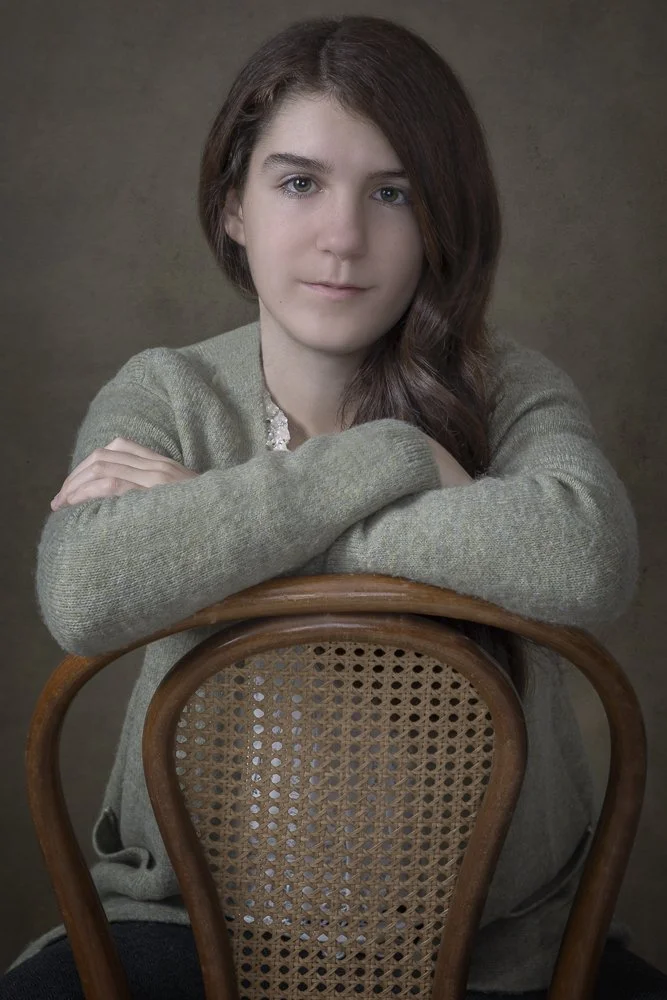 A young woman with long brown hair and fair skin sitting behind a wooden chair with a woven cane backrest, resting her arms on the chair's top rail, wearing a gray sweater, looking directly at the camera with a neutral expression.