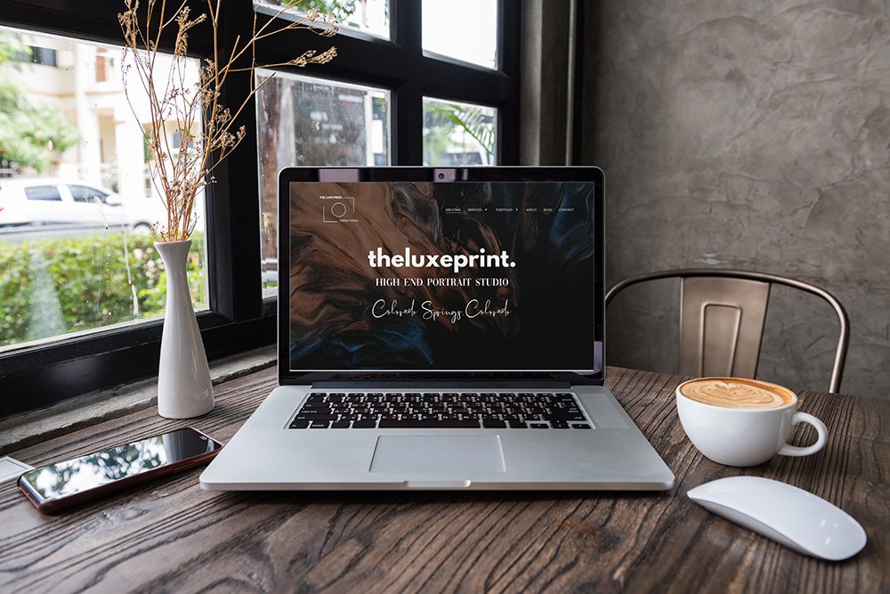 A laptop displaying 'theluxepprint. HIGH END PORTRAIT STUDIO' on a wooden table near a window, with a smartphone, a white vase with dried plants, a coffee cup with latte art, and a wireless mouse.