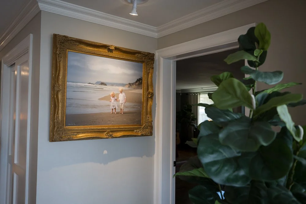 A framed photograph of two children holding hands on a beach hangs on a white wall in a living space. There are green houseplants to the right and a doorway leading to another room.