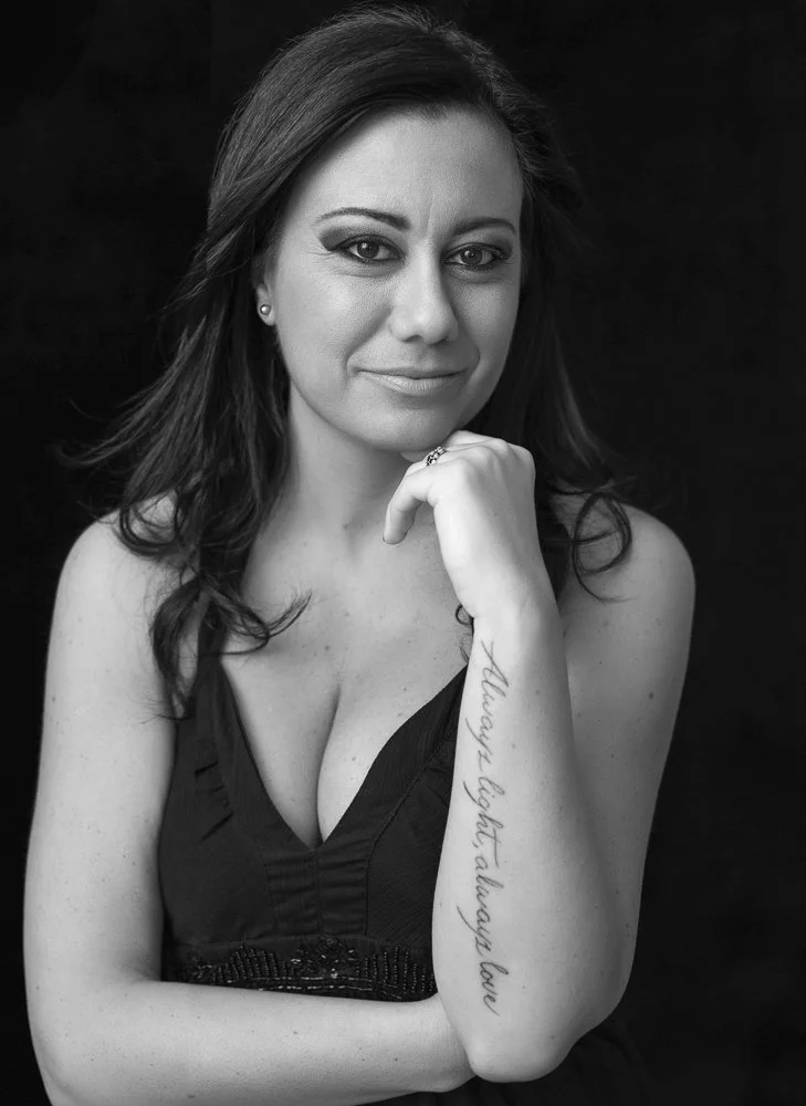 Black and white portrait of a woman with long dark hair, wearing a sleeveless top, resting her chin on her hand, with a tattoo on her forearm that reads 'Although right always love'.