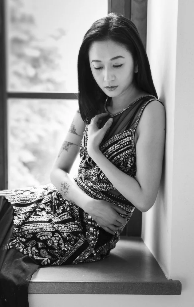 A woman with long dark hair sits by a window, looking down pensively, wearing a patterned sleeveless dress.