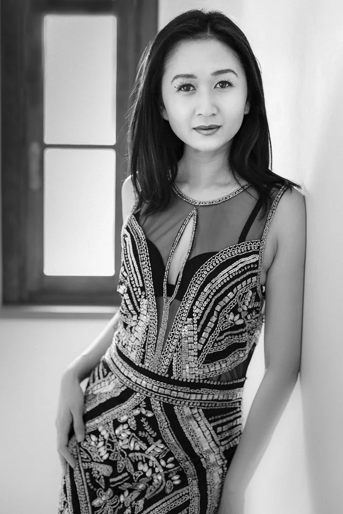 A woman with dark hair stands indoors near a wall, wearing a detailed, beaded dress with floral and geometric patterns, posing with one hand on her hip.
