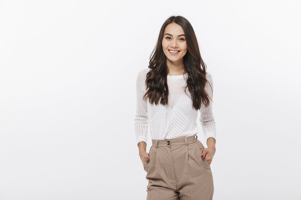 A young woman with long dark hair smiling, wearing a white sweater and beige pants, standing against a plain white background.