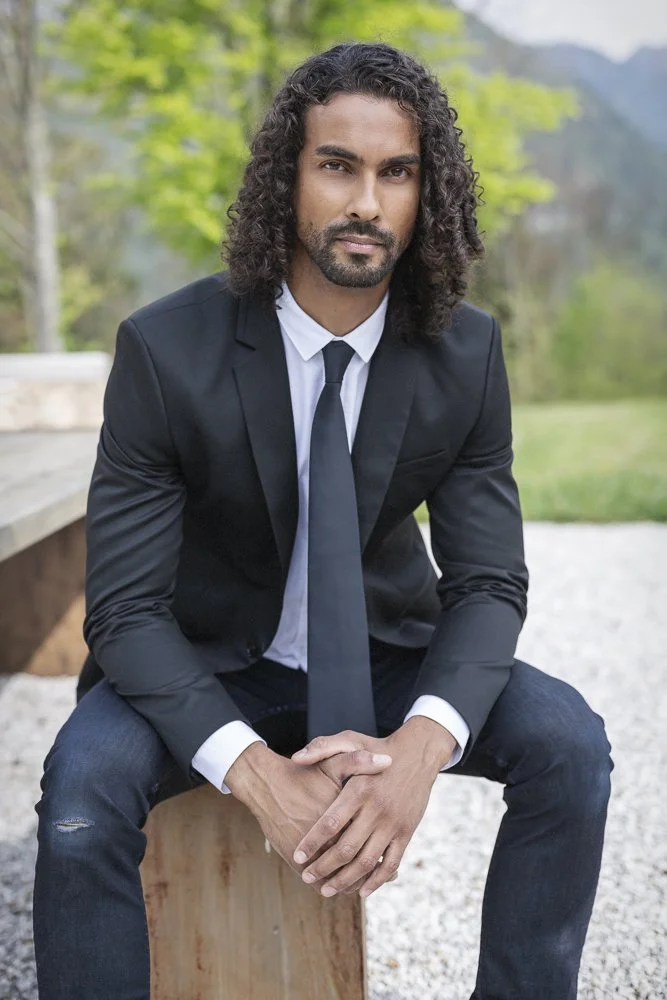 A man in a black suit, white shirt, and black tie sitting outdoors on a wooden bench, with greenery and trees in the background.