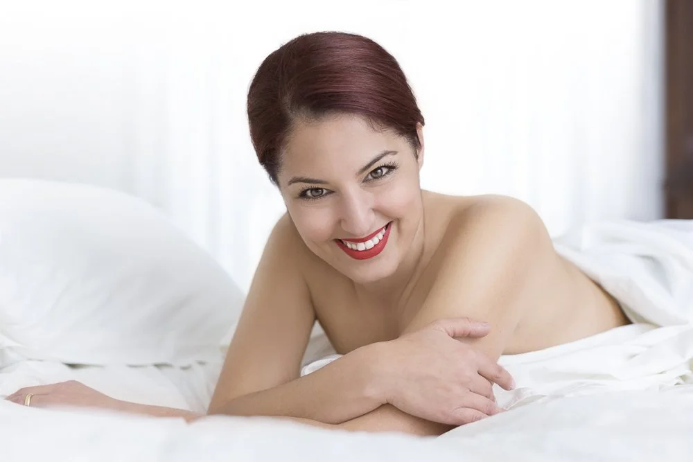 A woman with short red hair and red lipstick smiling while lying on a bed with white sheets in a bright room.
