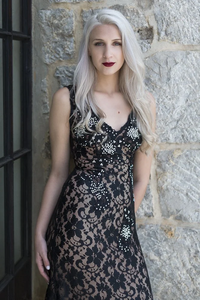 A woman with long platinum blonde hair wearing a black lace dress with floral and pearl embellishments standing against a stone wall.