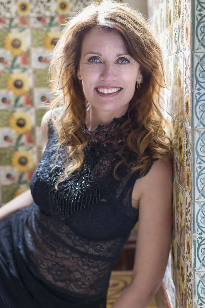 A woman with curly red hair and blue eyes is smiling and leaning against a colorful tiled wall.
