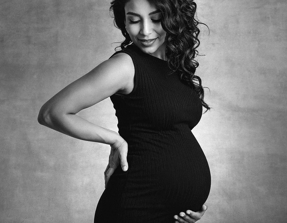 A pregnant woman with dark curly hair looking down, wearing a sleeveless black dress, standing against a textured background.