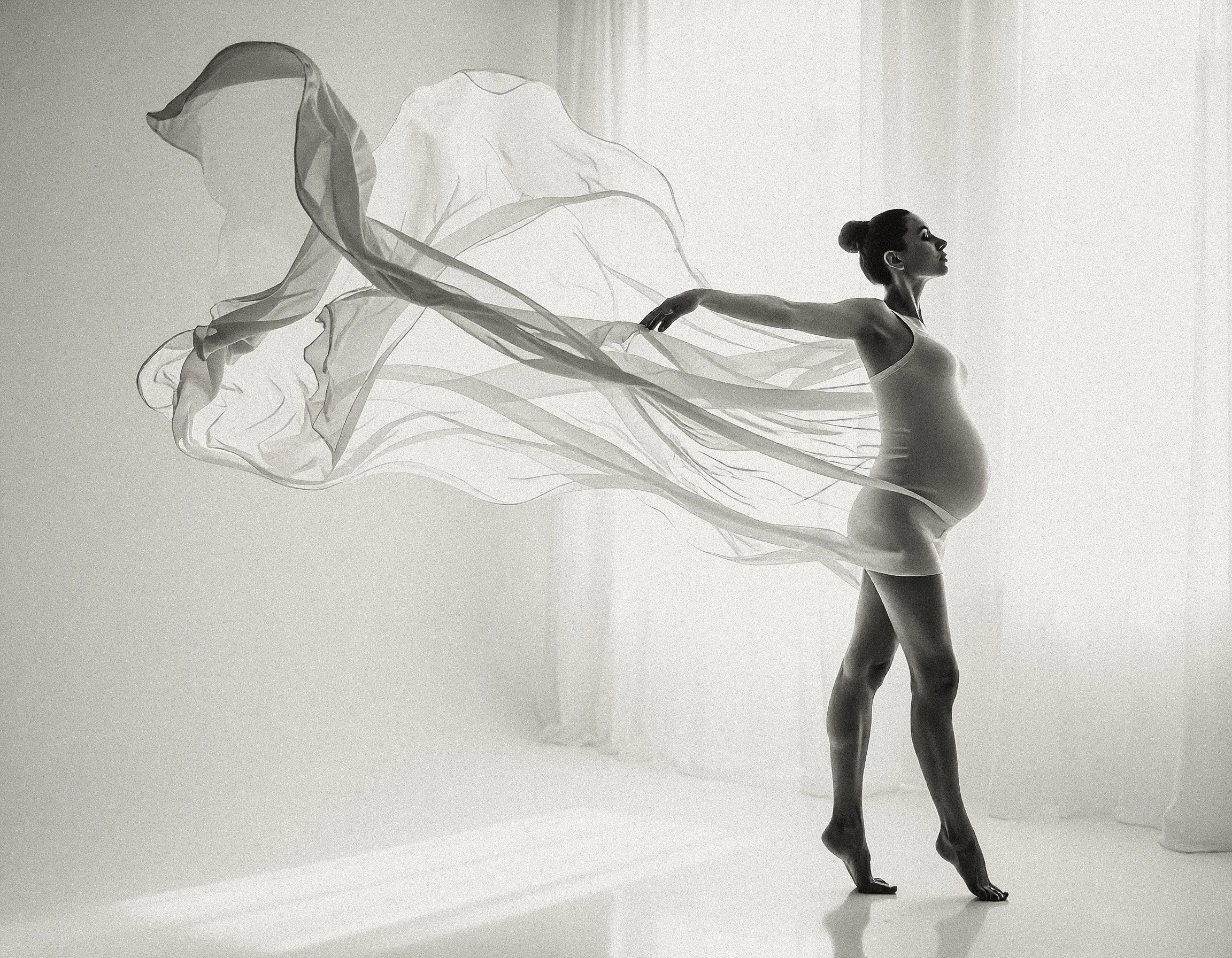 A pregnant woman dances gracefully in a minimalistic, softly lit room, with sheer fabric flowing behind her.