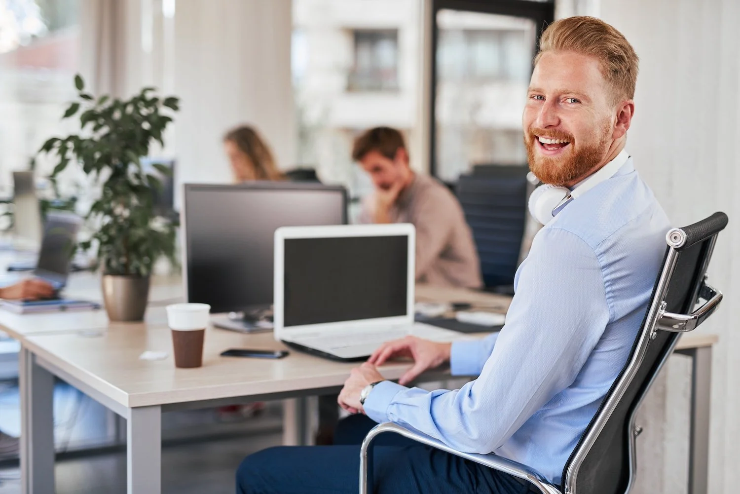 A bearded man with red hair smiling and sitting at a desk in an office, with two colleagues working at desks in the background.