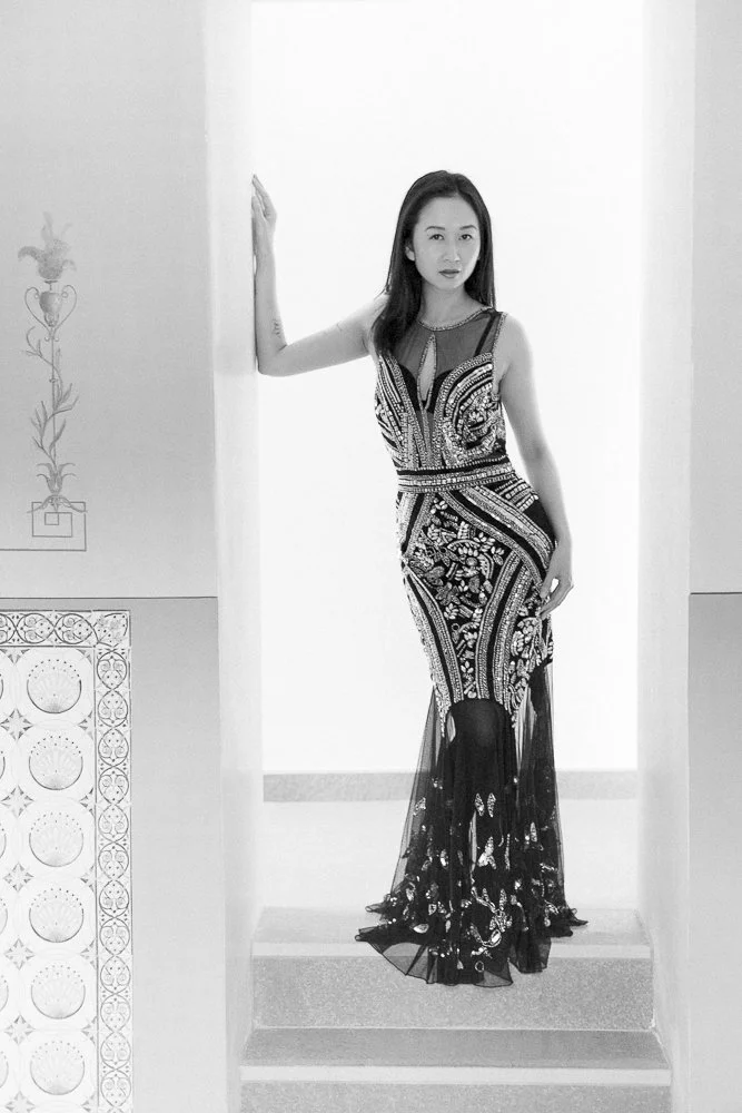 A woman in an elegant, sleeveless, embroidered black dress standing on stairs with her right arm raised, posing in a minimalist indoor setting.