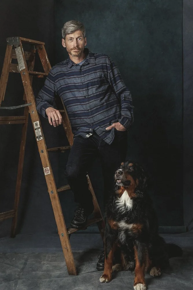 A man with gray hair and beard wearing a blue and gray plaid shirt standing next to a wooden ladder with a Bernese Mountain Dog sitting on the floor in front of him, against a dark background.
