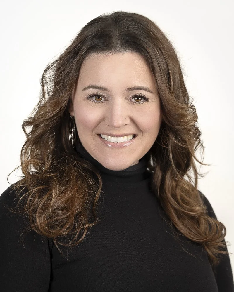 A woman with long, curly brown hair smiling and wearing a black turtleneck.