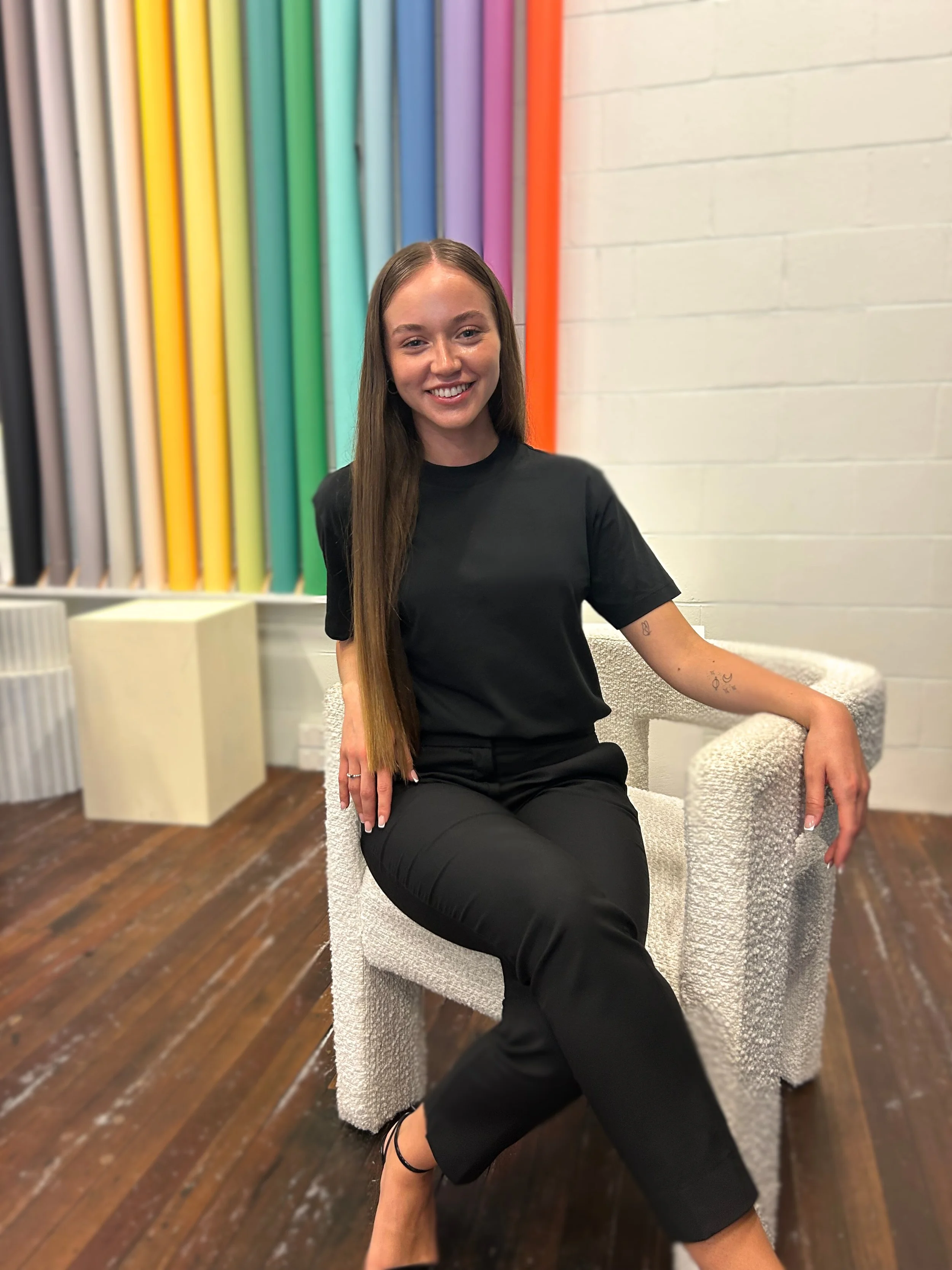 A young woman sitting on a textured white chair in front of a colorful wall display of large paint tubes, smiling at the camera.
