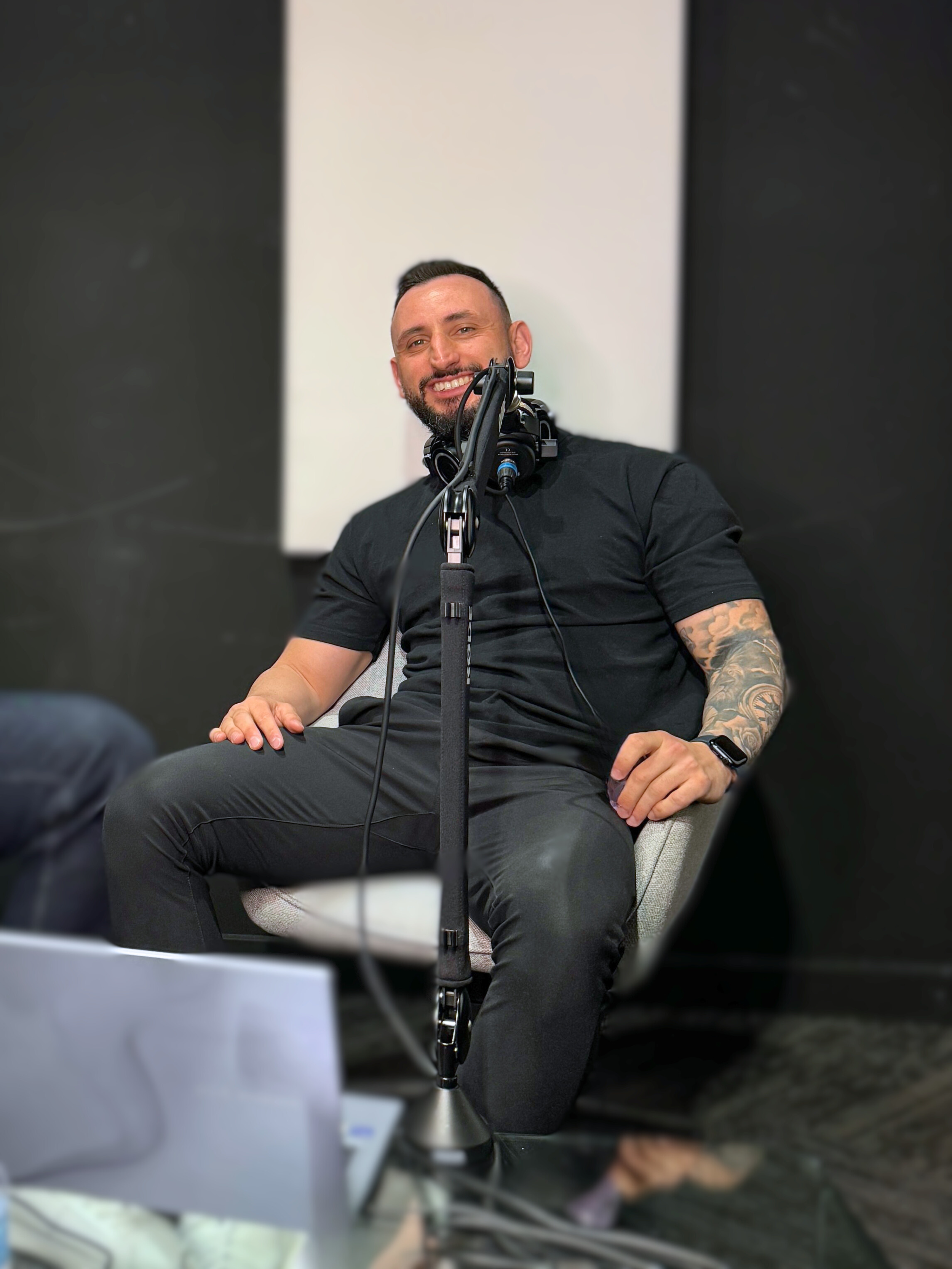 Smiling man with a beard, wearing a black t-shirt, sitting on a chair with headphones around his neck, in a recording studio or podcast setting.