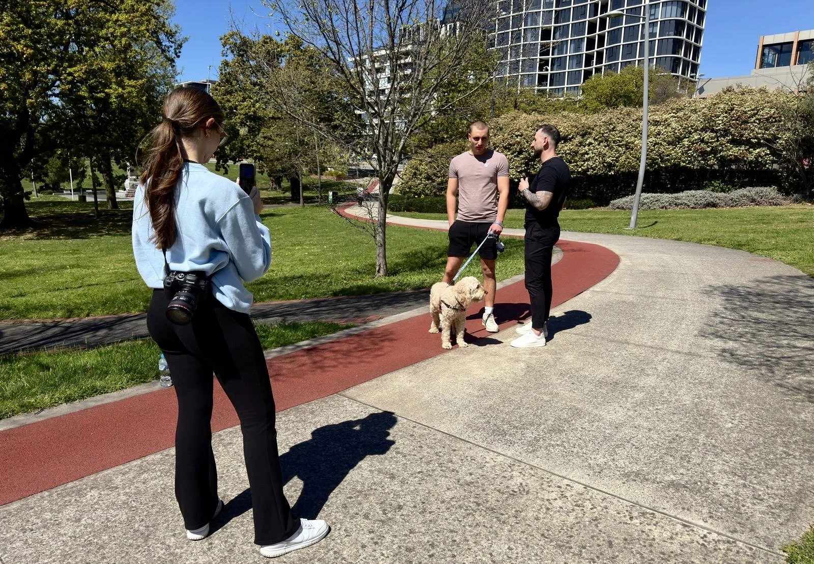 Two men and a woman with a camera around her neck are in a park. They are standing on a walking path, with one man holding a dog on a leash while they talk. The woman is taking a photo of the men with her phone. In the background, there are trees, shrubs, and modern buildings.