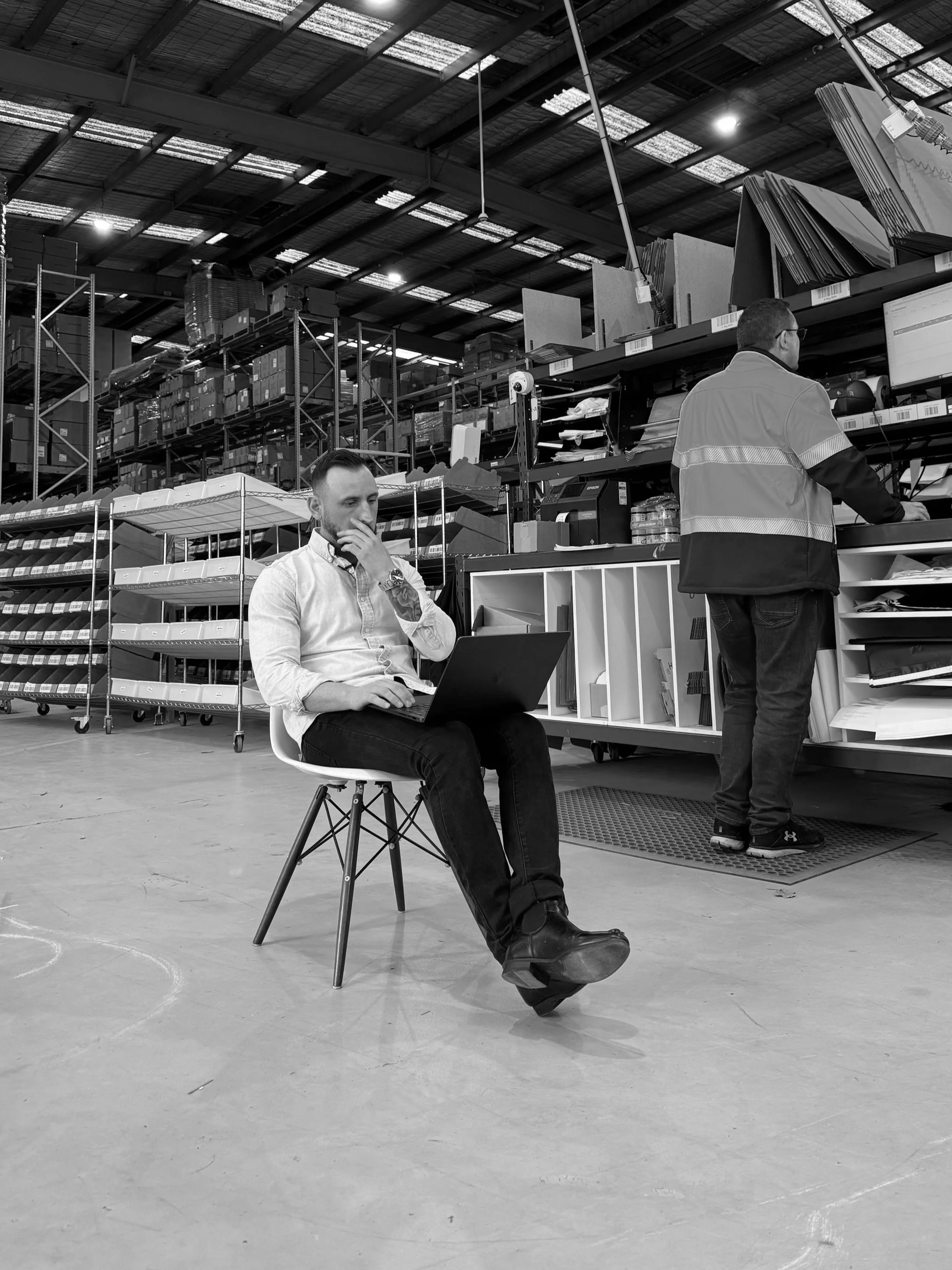 A man sitting on a chair with a laptop in a warehouse, while another man in a safety vest works at a shelf.
