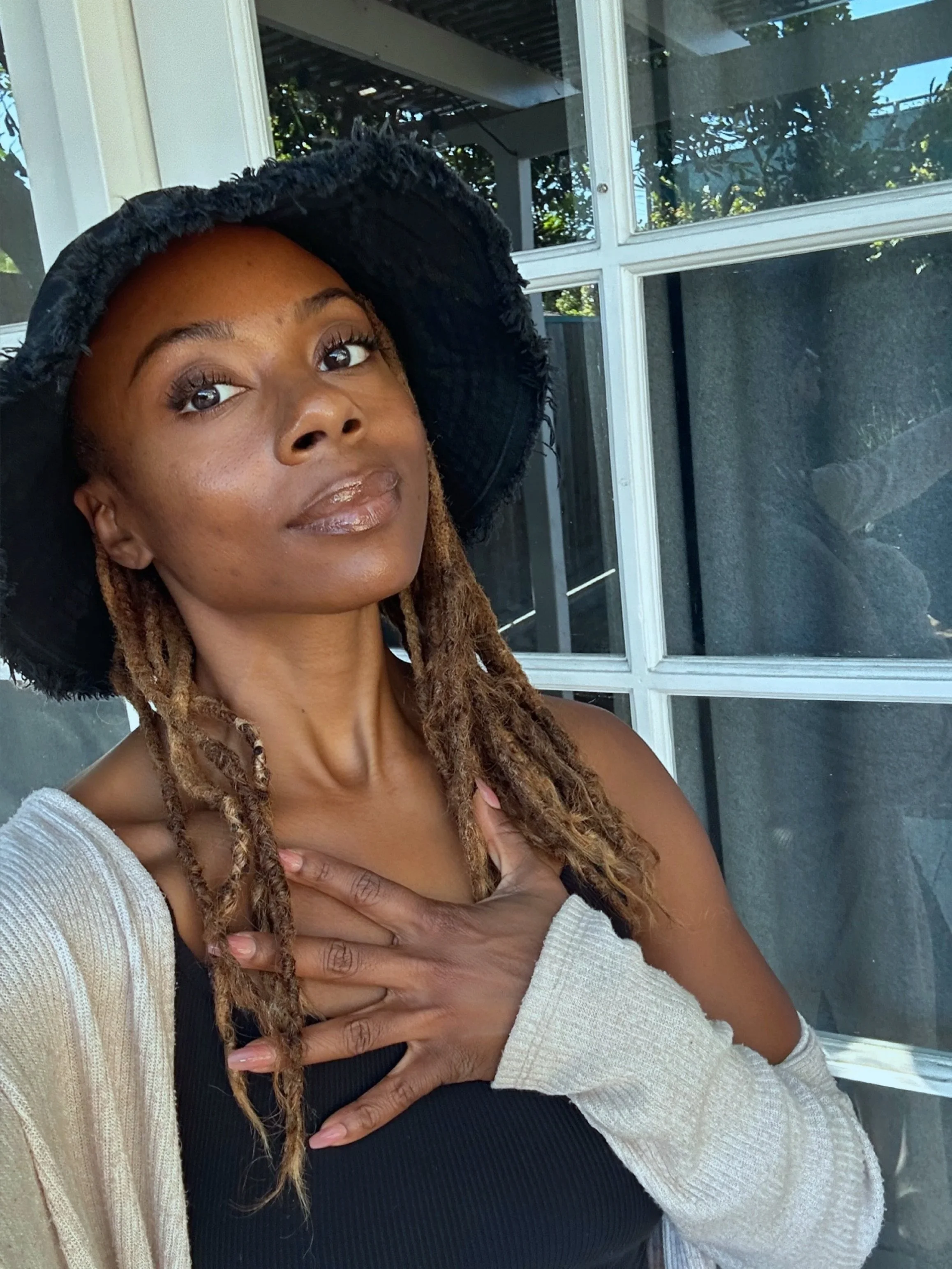 A woman with braided hair wearing a wide-brimmed black hat and a black top, posing with her hand on her chest in front of a window with a grid pattern and greenery outside.