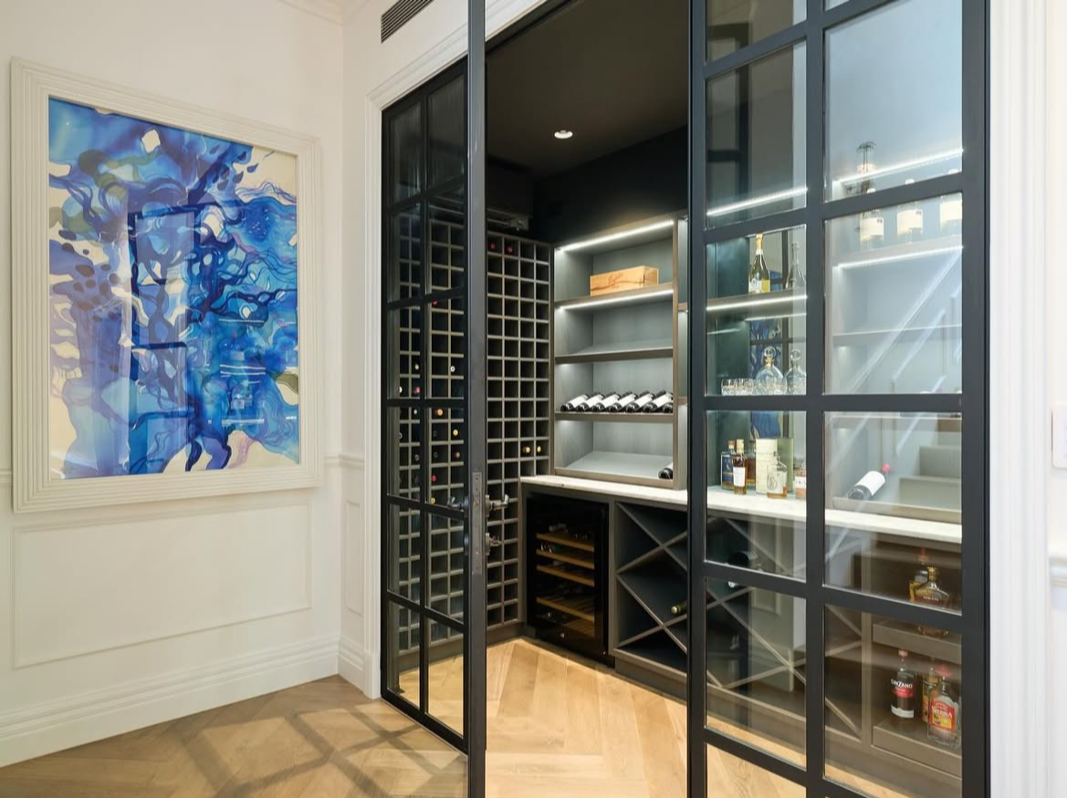 Modern wine cellar or bar area with glass-paneled black framing, dark shelves, and wine bottles, with an abstract blue and white artwork on the adjacent wall.