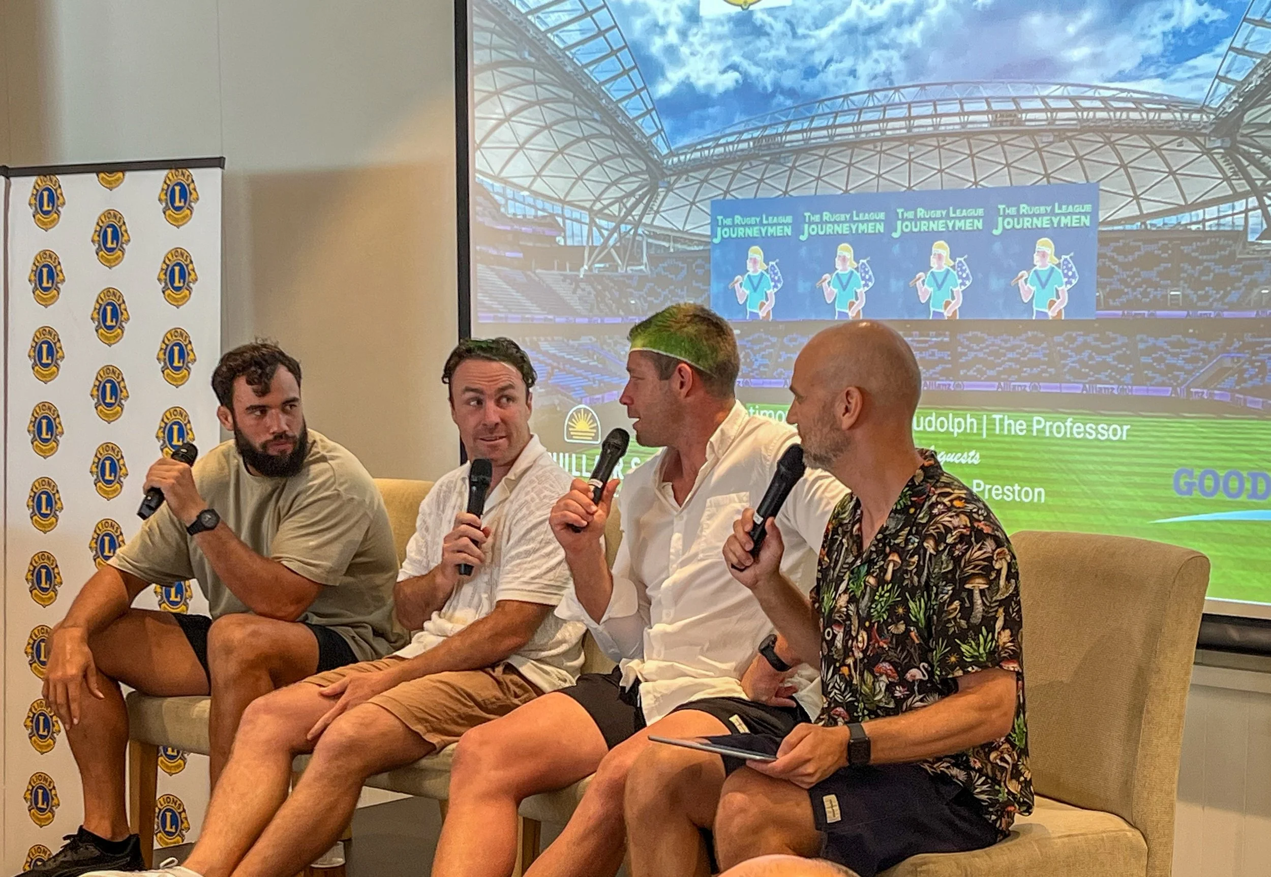 Four men sitting on a couch during a panel discussion, holding microphones, with a backdrop displaying a stadium and event branding.