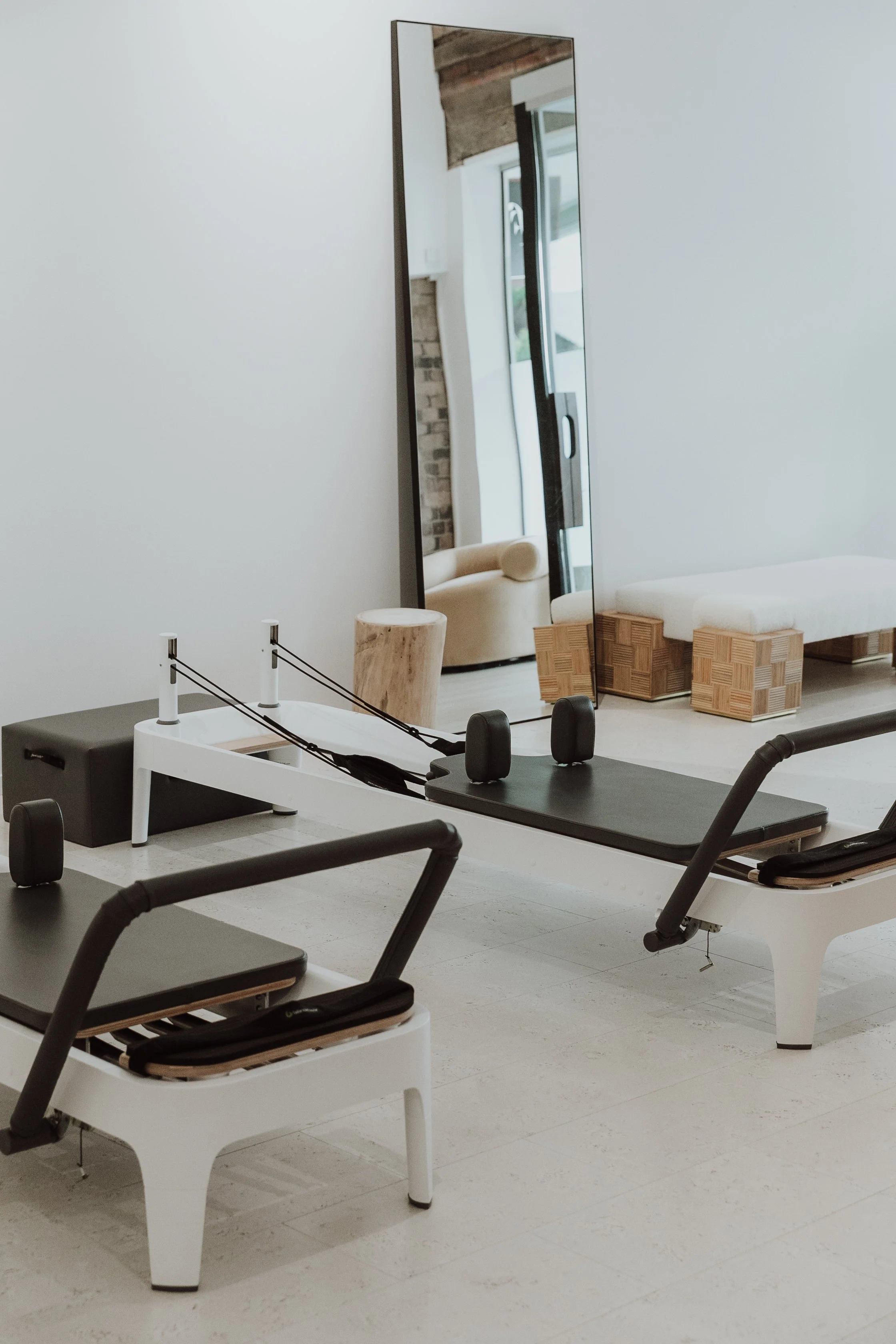 A modern, minimalistic fitness studio with Pilates reformer machines, a large floor mirror, and contemporary furniture in the background.
