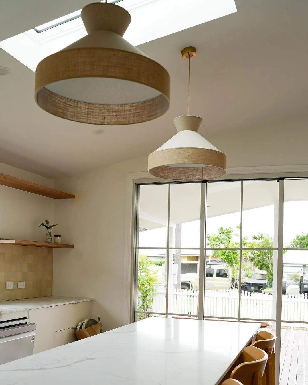 Modern kitchen with white countertops, wooden chairs, large sliding glass doors, hanging lamps with white lamp shades and wooden accents, and a view of a porch and parked cars outside.