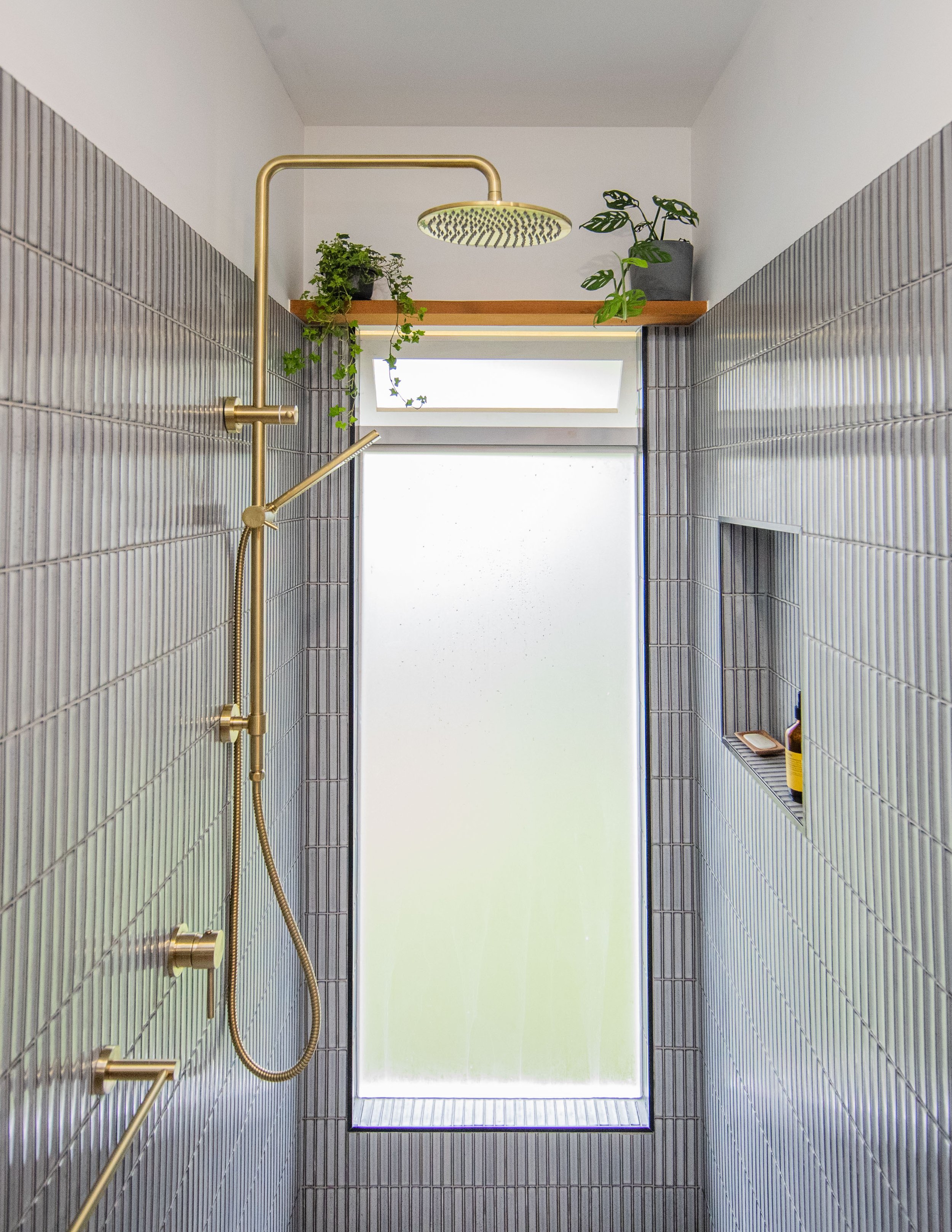 Modern shower with gold fixtures, gray tiled walls, built-in niche with toiletries, frosted glass window, and a wooden shelf with potted plants.