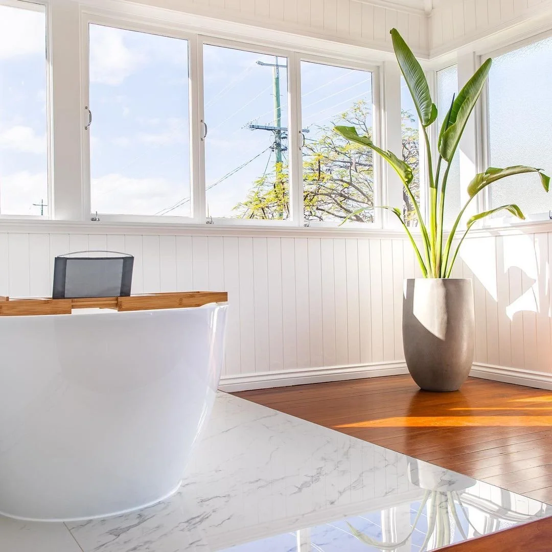 Bright bathroom with a white bathtub, large windows, a wooden floor, a tall potted plant, and white paneling on the walls.