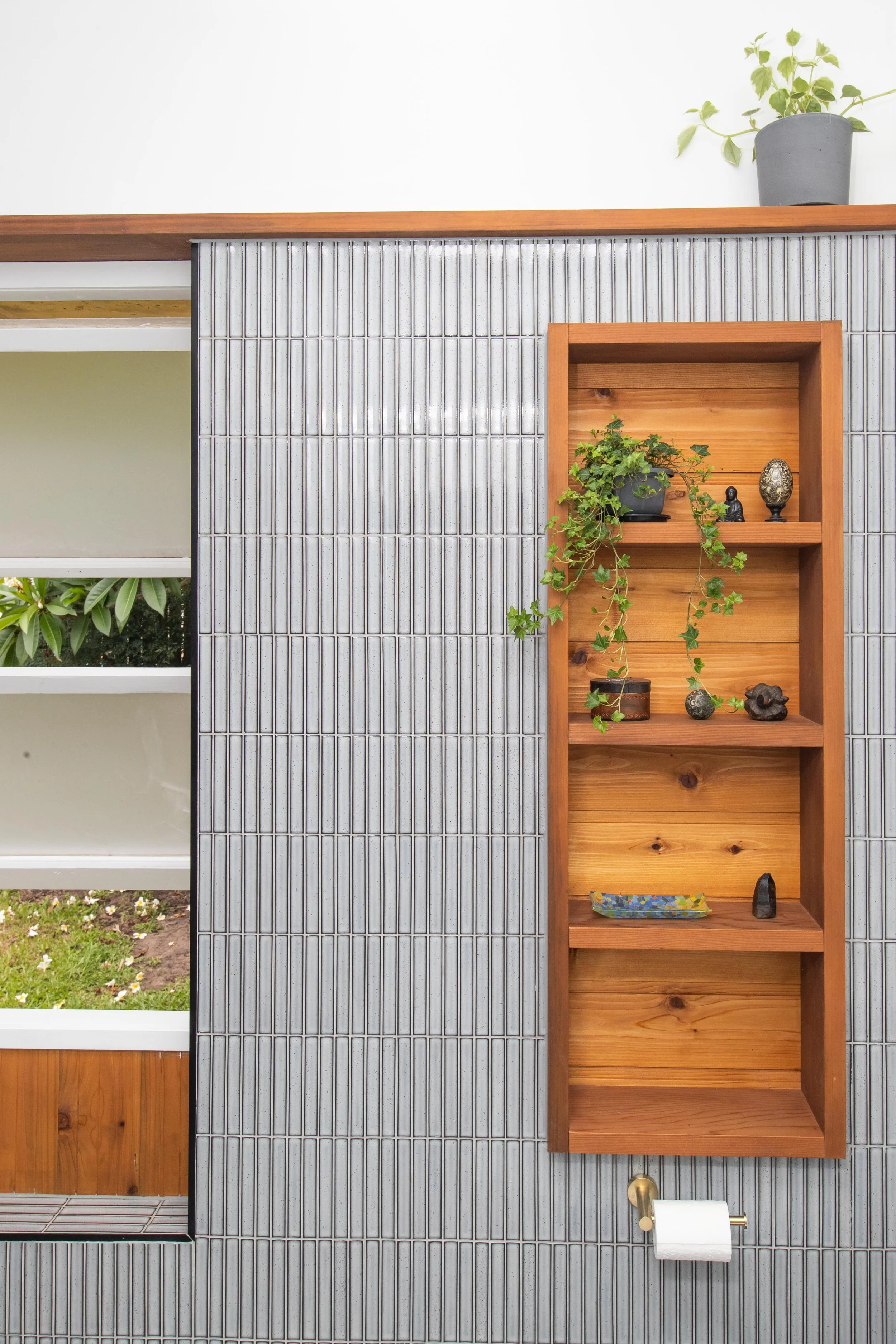 Decorative wooden shelving unit with small plants and ornaments on a tiled wall, with a potted plant on top and a toilet paper holder below.