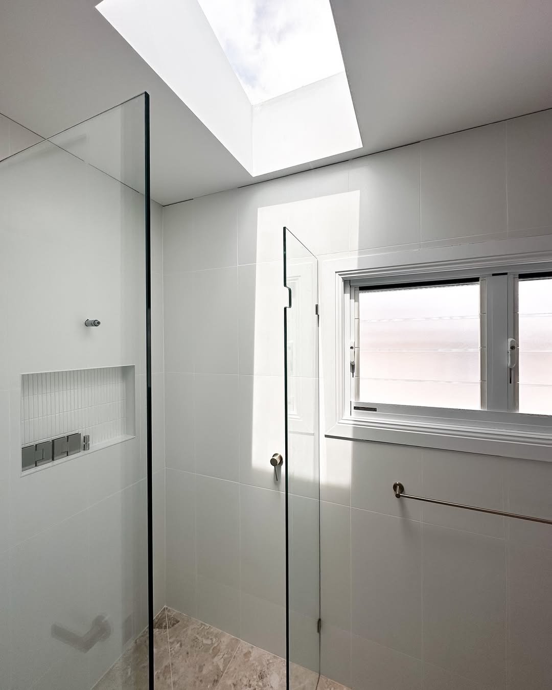 A modern bathroom with white tiled walls, a glass shower enclosure, a skylight above, and a window on the wall, with a towel bar underneath.