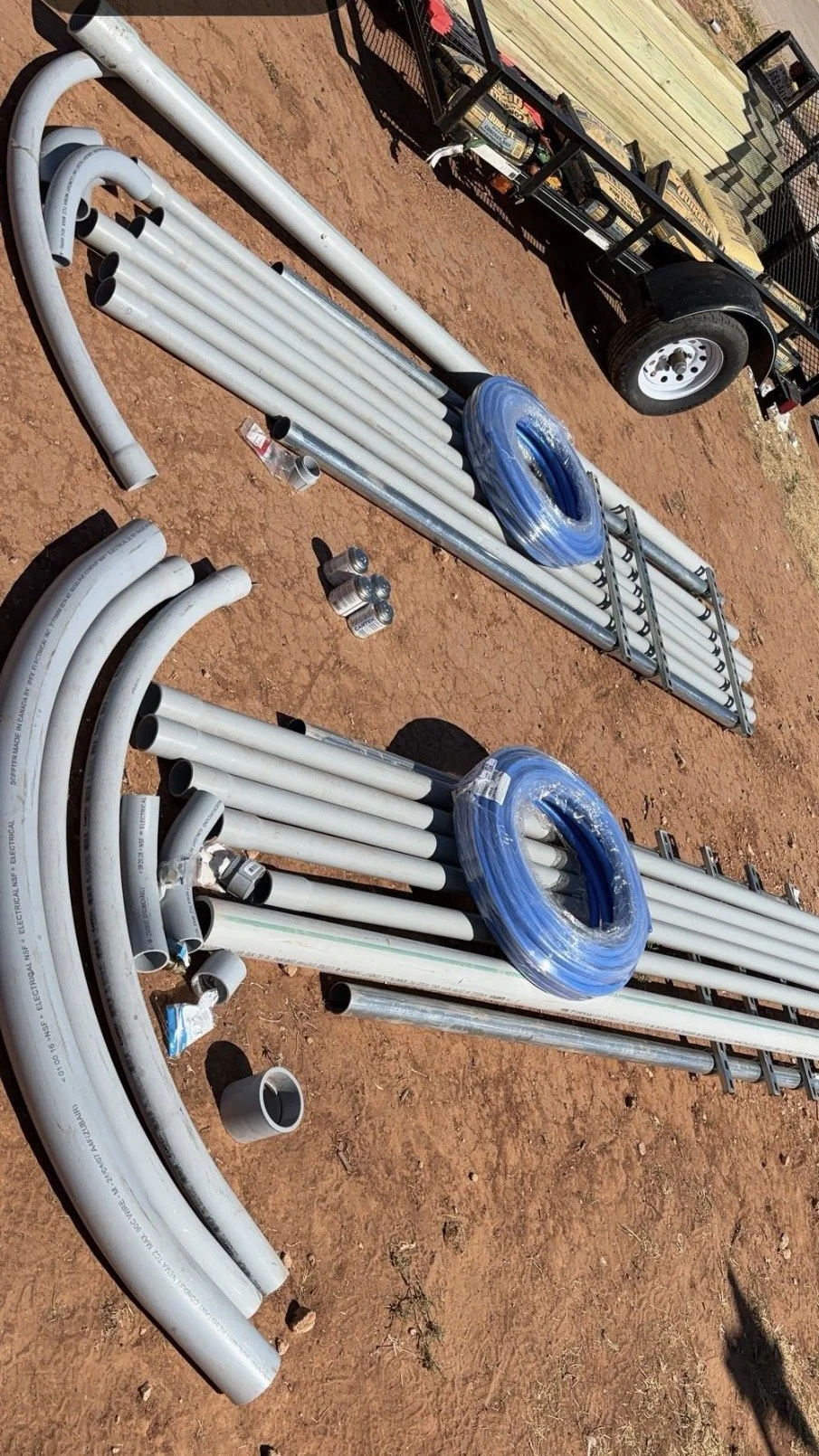 PVC pipes, tubing, and fittings laid out on reddish dirt ground, with a metal utility cart and a trailer in the background.