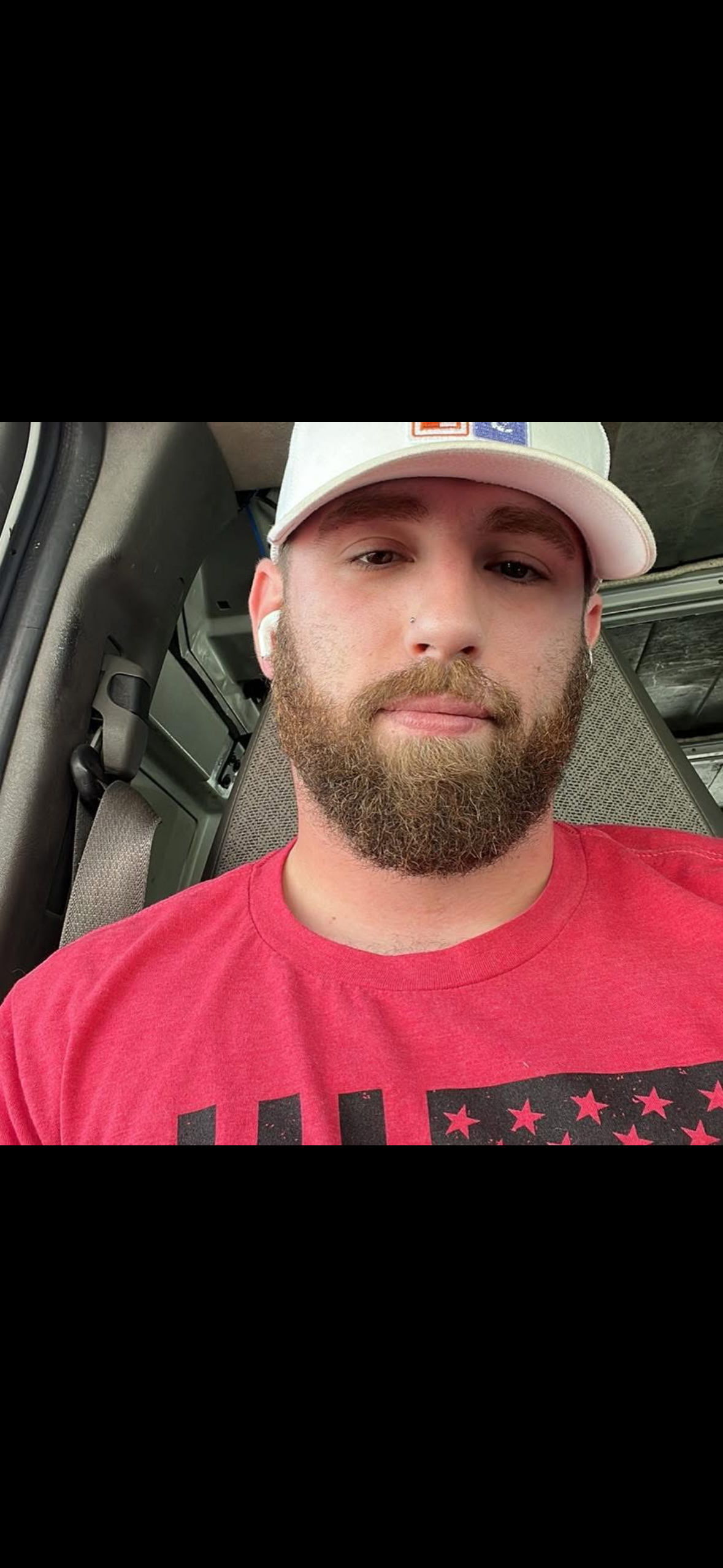 A young man with a beard wearing a white cap, red T-shirt, and wireless earbuds inside a vehicle with a gray interior.