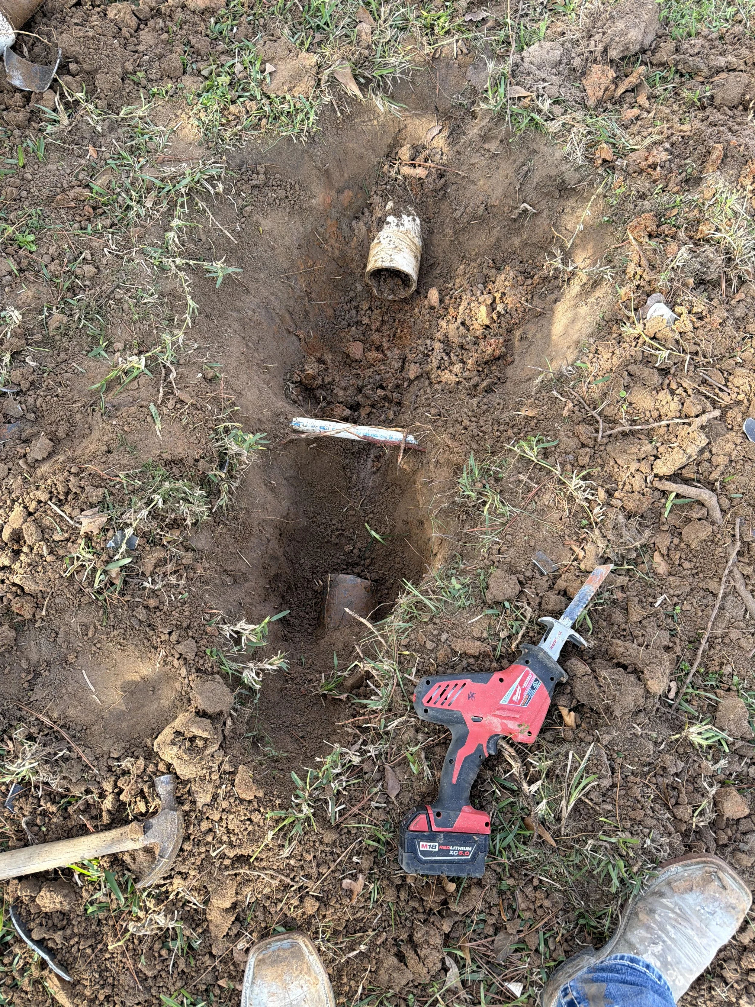 A partially dug trench with a pipe in the ground, a handheld reciprocating saw, a hammer, and shoes visible, indicating ongoing pipe repair or installation.
