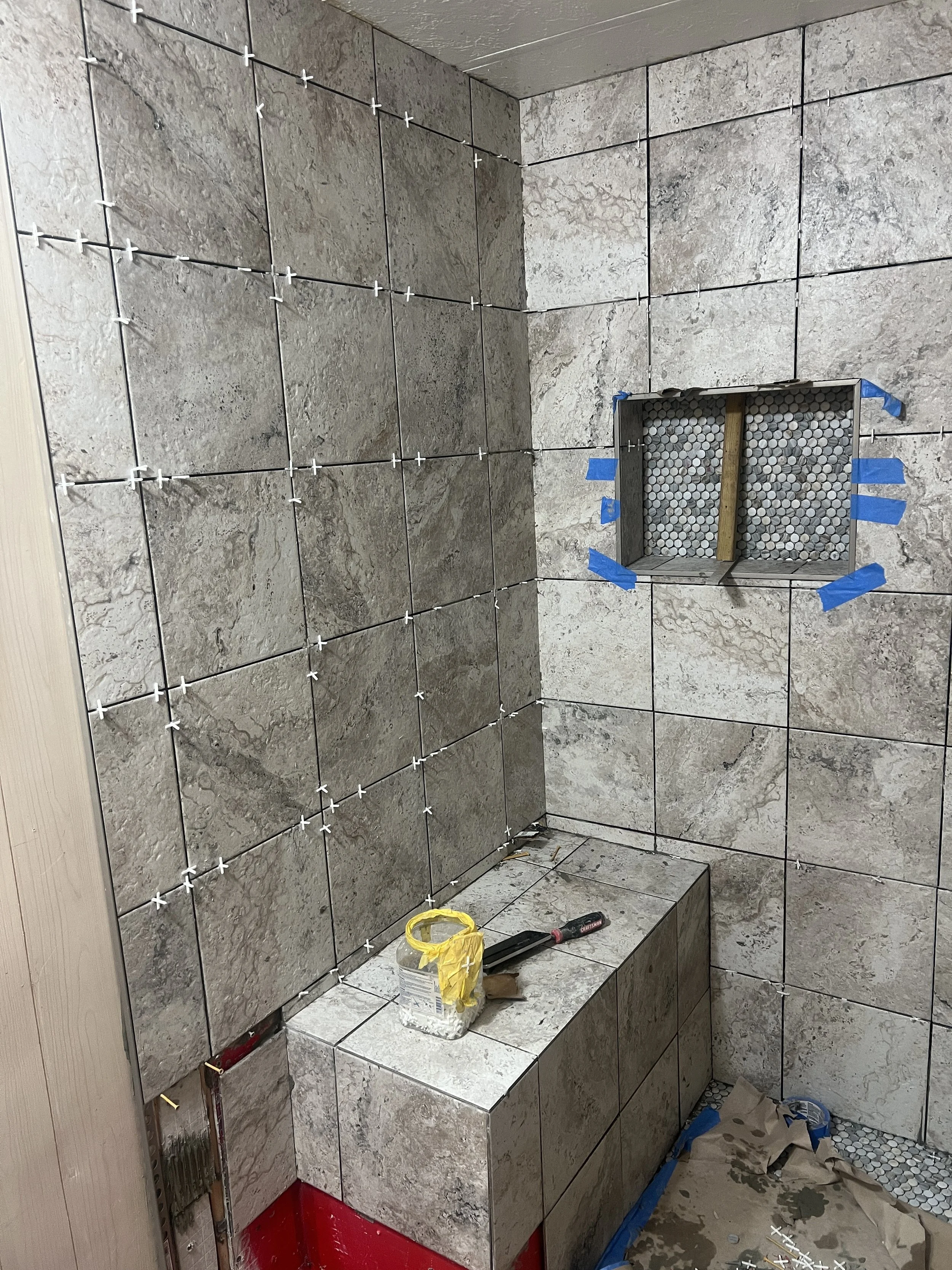 Bathroom wall with gray tiles being installed, a cut-out for a niche, and tools on a ledge.