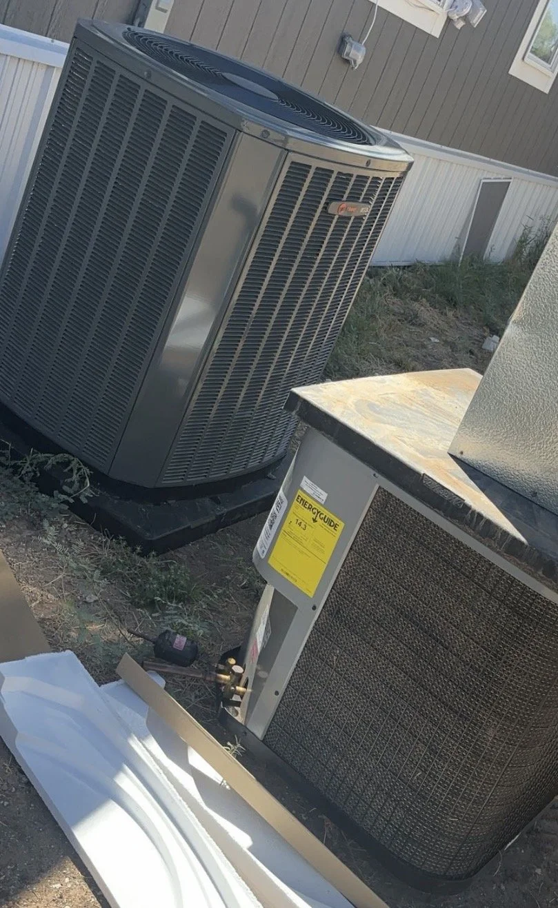 An outdoor view showing a large air conditioning unit and a hot water heater placed on the ground near a house with brown siding and a white fence.
