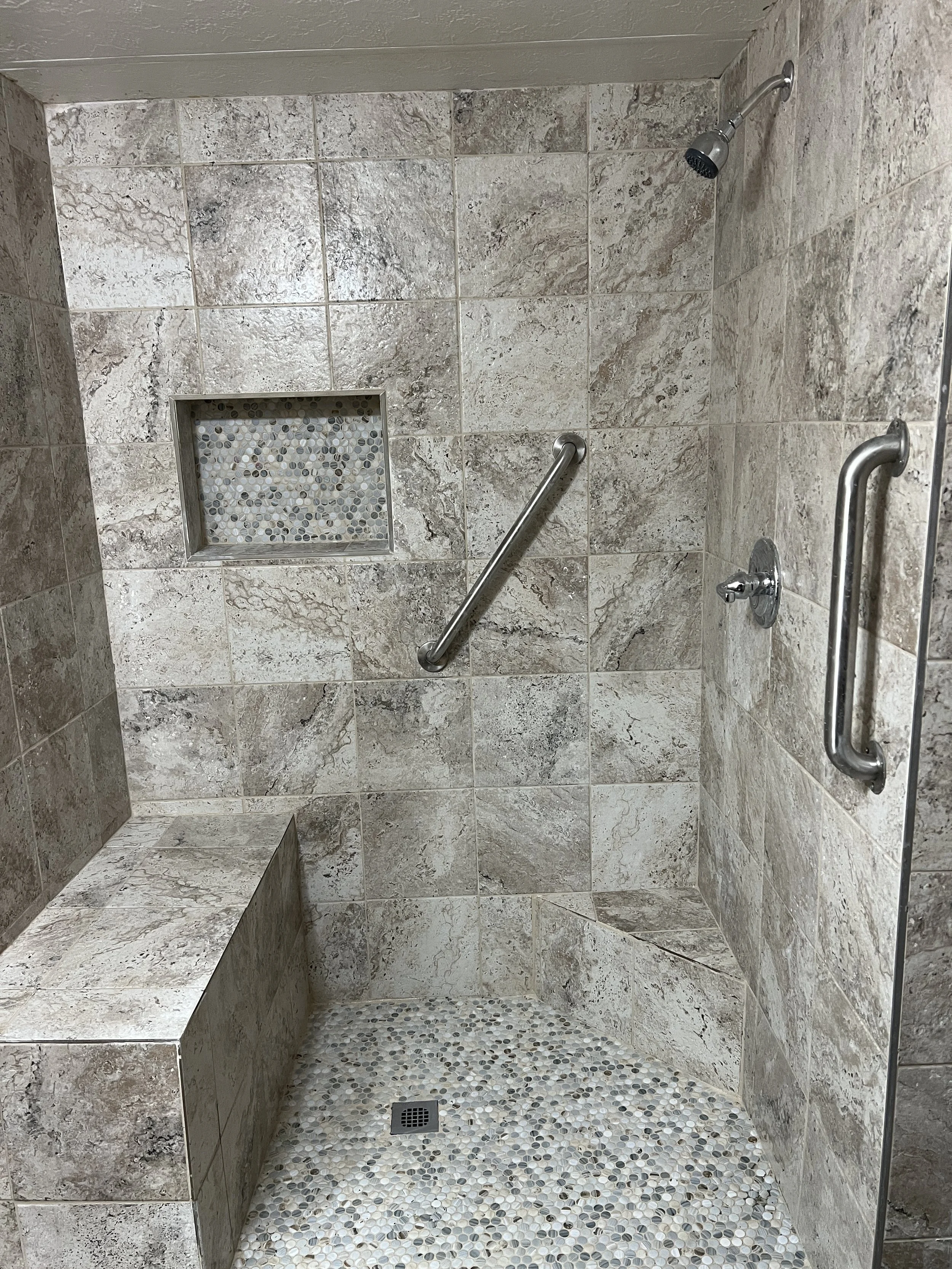 Shower with beige and brown marble tiles, a built-in bench, metal grab bars, a rainfall shower head, a small tile niche, and a pebble floor drain.