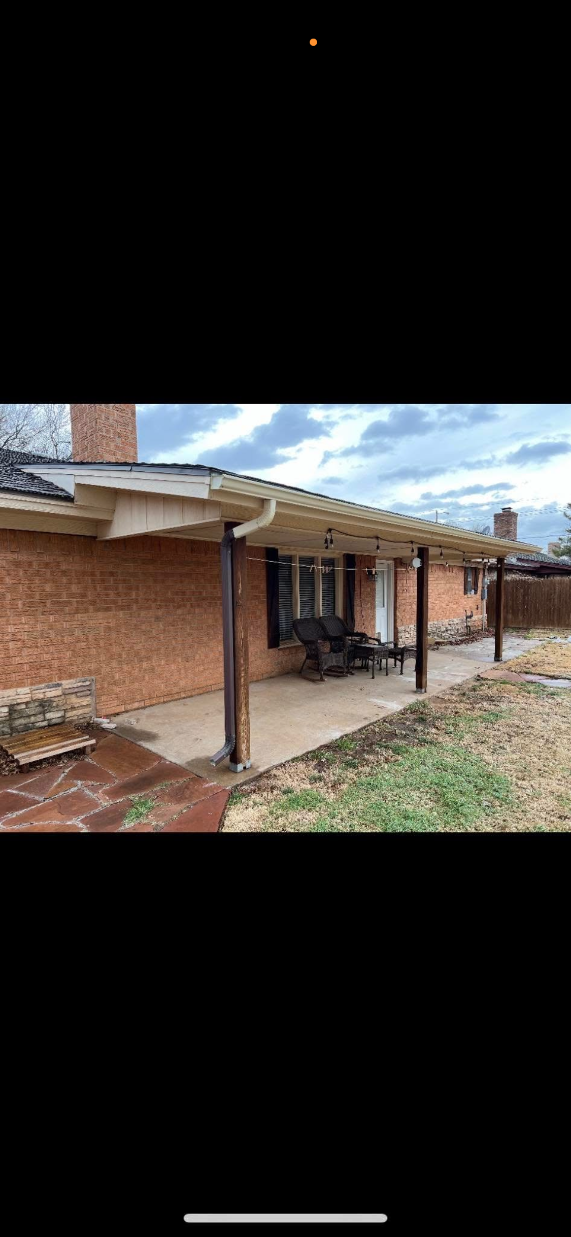 Backyard patio with outdoor furniture, brick house, wooden support posts, and cloudy sky.