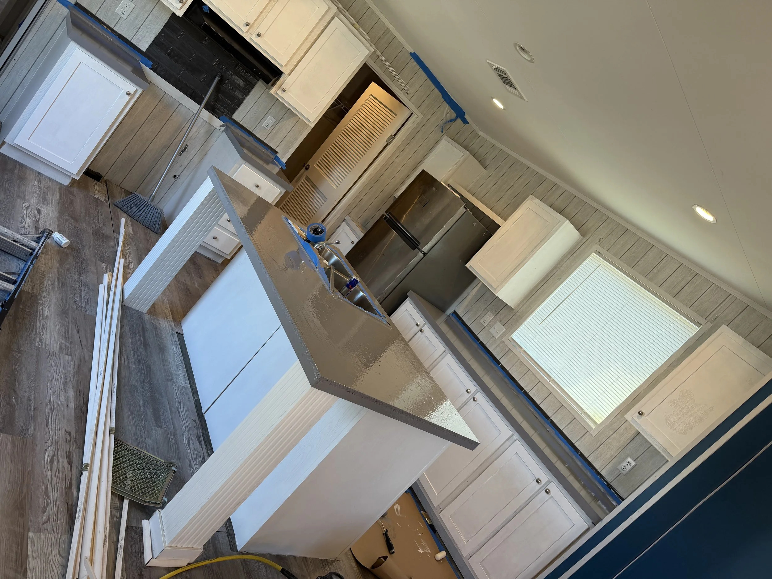 Kitchen under renovation with white cabinets, a large island with a gray countertop, and a stainless steel refrigerator. Construction tools and materials are scattered around, and the walls have a wood paneling finish.