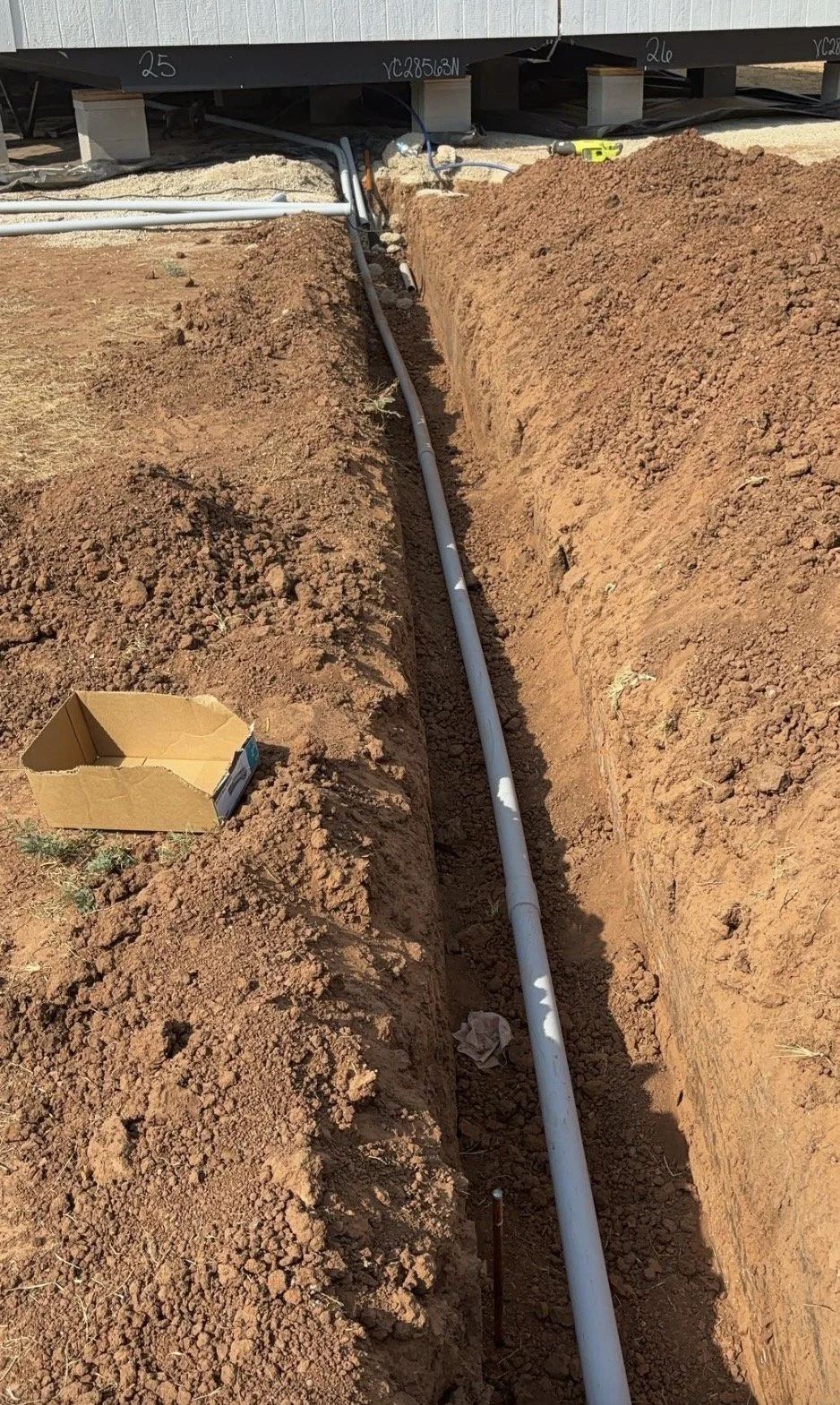 A trench with a white pipe installed along its length, with construction tools and equipment visible nearby, under a building raised on blocks.